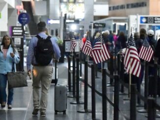 Travelers with luggage walk through an airport terminal lined with small American flags near a security checkpoint.