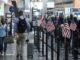 Travelers with luggage walk through an airport terminal lined with small American flags near a security checkpoint.