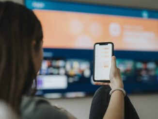 A girl uses a smartphone while watching a TV mounted on a living room wall.