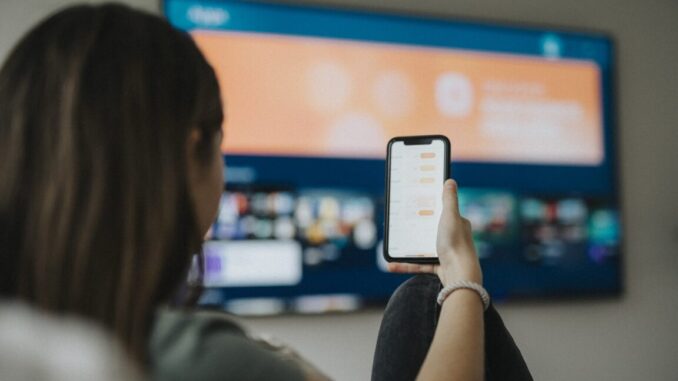 A girl uses a smartphone while watching a TV mounted on a living room wall.