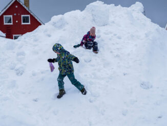 Two children in winter clothing play on a large mound of snow in front of red houses.