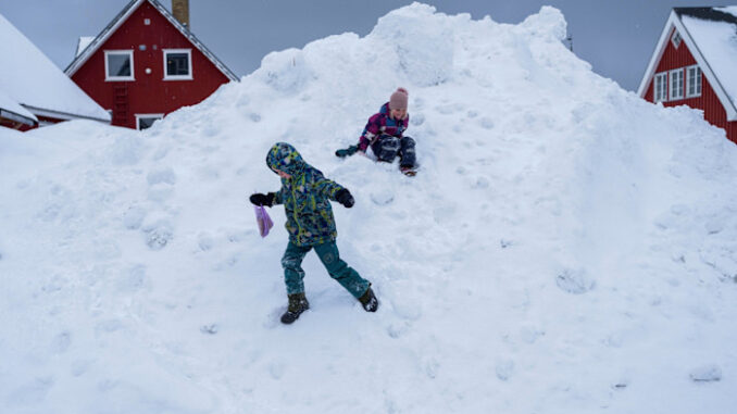 Two children in winter clothing play on a large mound of snow in front of red houses.