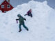 Two children in winter clothing play on a large mound of snow in front of red houses.