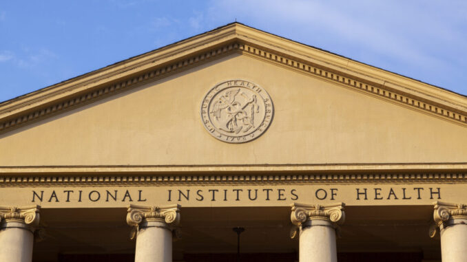 A view of the exterior of a building with a triangular top supported by pillars. Beneath a circular logo are the words "national Institutes of Health"